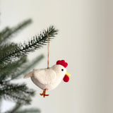 A handcrafted felt chicken ornament is gracefully displayed on a Christmas tree branch, set against a soft white backdrop. This charming white felt chicken features a vibrant red comb, a bright yellow beak, and distinct orange legs. A jute string is attached for easy hanging, highlighting its unique artisanal quality.