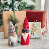 Two handcrafted felt Christmas gnome ornaments rest on a reflective marble surface, in front of a blurred twinkling Christmas tree and wrapped gifts, creating a warm holiday scene. One grey felt Christmas gnome features a textured body, red pom poms on its hat, and a long white beard. The other red felt gnome displays intricate white detailing on its hat and a fluffy grey accent above its long white beard.
