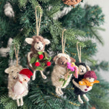 Three unique felt sheep Christmas ornaments are suspended from the branches of a green Christmas tree adorned with pine cones, creating a joyful and festive atmosphere. Each handcrafted felt sheep ornament is suspended by a jute string. One brown sheep wears a green wreath with red berries, a white sheep dons a red Santa hat, and a cream sheep holds a bouquet of pink, yellow, and green flowers.
