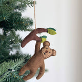 A brown felt monkey Christmas ornament hangs playfully from a branch on a Christmas tree, set against a plain white wall. This handcrafted felt monkey ornament features a cheerful stitched face, depicted swinging from a brown branch with green leaves at each end. A jute string is attached to the branch, allowing it to dangle charmingly amidst the tree's green needles.