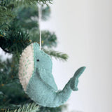 A serene wool felt whale Christmas ornament hangs from the green needles of a Christmas tree, adding a touch of handmade charm. The gentle whale has a light blue body, a creamy white head section, and is detailed with light green stitches. A dark eye and a curved tail complete its whimsical design, highlighting its soft, textured appearance.