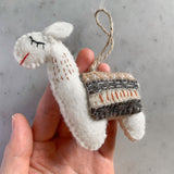 A female hand gently holds one white felt llama Christmas ornament against a mottled gray and white surface. This handcrafted felt ornament features a jute string loop for hanging. The white llama has a rectangular saddle with brown, gray, and white stripes accented with red. Its face shows closed black eyes and a red stitched mouth, with small red stitches highlighting its body.