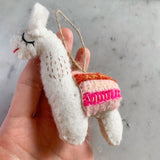 A wool felt llama ornament is held in a female hand against a soft grey background. This charming white llama, crafted from textured wool felt, features closed embroidered eyes, a small red mouth, and a colorful saddle with pink, orange, and cream striped embroidery. A jute string is attached, ready for hanging this unique Christmas tree ornament.