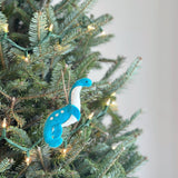 A felt dinosaur ornament hangs on a green Christmas tree adorned with warm white lights, creating a festive atmosphere. This unique felt Christmas ornament features a long-necked dinosaur made of blue and white felt. Its body is blue with white dots, and its neck is white with a blue head. It hangs from a jute string, blending with the tree's rustic charm.