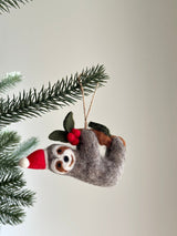 A felt Christmas sloth ornament hangs from a Christmas tree branch. This charming felted sloth wears a red Santa hat with a white pompom and holds a small brown felt branch adorned with green leaves and red berries. The handcrafted ornament hangs by a jute string.