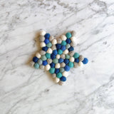 A set of four blue star felt coasters is displayed on a marbled white surface. These unique, handmade felt coasters are crafted from hand-stitched half-inch felt balls in shades of blue, grey, teal, and white, creating a textured, layered effect.