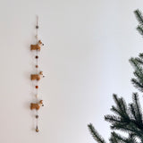 A charming Felt Corgi Garland is displayed against a clean white wall, next to a green pine tree branch. The playful garland features three light brown felt corgi figures with white faces and paws, spaced along a string. Between each corgi are felt balls in gray, cream, and brown. A copper bell dangles at the bottom, adding a simple decorative touch.