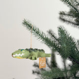 A needle felted crawling gator ornament hangs playfully on a lush green Christmas tree against a plain white background. This green gator ornament is intricately detailed with a visible back, eyes, a pink nose, and white teeth. Made of felted wool, a brown jute thread is attached for easy hanging.