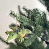 A felt crawling gator ornament hangs playfully on a green Christmas tree, set against a blurred bright background. This cheerful wool felt gator features a green body with darker green markings, small green eyes, a pointy snout with white felted teeth, and four short legs. A jute string attached at the top allows it to hang from the branch.