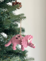 A pink triceratops felt dinosaur ornament hangs on a vibrant green Christmas tree. This unique Christmas ornament, crafted from wool felt, features brown and white spots, a white horn, and a red smiling mouth. A jute string is visible for hanging, with lush green pine needles and a pine cone in the background, creating a festive scene.