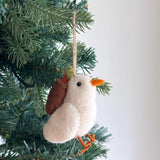 A charming wool felt dove ornament hangs from a green Christmas tree branch. This pure white felt dove features a small black eye, an orange beak, and orange felt feet perched on a needle. A jute string is attached for hanging, as the dove gracefully carries a delicate olive twig.