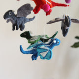 A colorful felt dragon baby mobile, featuring several handcrafted felt dragons, is displayed in a bright, cheerful nursery setting. The mobile includes felt dragons in red, grey, green, and blue, each with small wings and delicate details. Each dragon is suspended by a thin white string, creating a floating effect, and interspersed with bright pink felt balls. Its whimsical and comforting design is perfect for a baby's nursery.