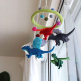 A felt dragon baby mobile, featuring colorful felt balls and dragons, is displayed against a bright window, creating a whimsical and airy atmosphere. The mobile has a light green felt ring from which four blue, red, gray, and green felt dragons, detailed with wings and tails, are suspended. Pink, light blue, gray, and olive green felt balls also hang, all attached with thin white strings.