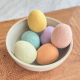 A set of six handcrafted wool felt Easter eggs rests in a speckled ceramic bowl on a woven surface. The life-sized eggs, in pastel macaron-inspired shades of yellow, light green, light blue, orange, lavender, and pink, are grouped closely, exuding a soft, inviting charm. The natural textures enhance the cozy, artisanal presentation.