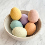 Handcrafted felt Easter egg ornaments are nestled in a speckled ceramic bowl, set against a bright white marble background. These life-sized, uniformly egg-shaped felt ornaments feature a soft, textured 100% wool felt surface. They come in an array of pastel colors including yellow, pink, lavender, light green, light blue, and peach, evoking a gentle springtime charm.