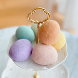 Six life-sized wool felt Easter eggs are elegantly displayed on the top tier of a white, two-tiered decorative stand with a gold handle. The pastel felt Easter eggs include shades of light blue, peach, yellow, pink, green, and lavender. They are densely arranged, filling the stand's top tier, which features a delicate, floral-patterned rim, evoking a dreamy springtime charm.