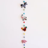 A playful felt farm animal garland hangs vertically against a plain white background. It features a grey wool felt horse with a black mane, a white cow with black spots and a pink snout, a rooster with a red head and tail feathers, and a pink pig. Colorful felt balls are strung between the needle-felted animals, with a single copper bell at the bottom.