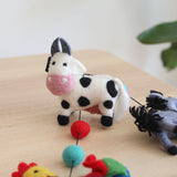 A wool felt farmland animal garland is displayed on a light brown wooden surface. This charming handmade garland features 3D needle-felted animals, including a white milk cow with black spots, a pink snout, and gray ears, a gray horse, and a rooster. The figures are suspended by black strings, ending with a copper bell. A blurred green plant is in the background.