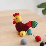 A colorful wool felt rooster, part of a Felt Farmland Animal Garland, rests on a light brown wooden table against a simple white background with a hint of green. This 3D needle-felted rooster features a red comb, yellow head, maroon body, and a vibrant tail in red, green, and blue.