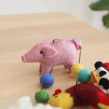 A handcrafted wool felt farmland animal garland rests on a light wooden surface, creating a calm and inviting atmosphere against a soft, blurred green background. The garland features vibrant, 3D needle-felted animals: a pink pig with a curled tail, a black and white cow, a red rooster with a yellow beak, a white sheep, and a horse. A small copper bell is attached at one end. This charming garland offers a playful aesthetic.