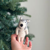 An Alaskan Husky Dog felt finger puppet is held by a female hand against a festive Christmas tree. This handcrafted wool felt finger puppet is gray and white, featuring intricate needle felt details of the Husky's erect triangular ears and thick double coat.