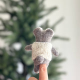 A handcrafted wool felt baby koala finger puppet is gently held in a female hand, poised for interactive play against a softly blurred background. This charming koala features soft gray fur, tiny ears, and a small tail, with visible needle-felted details. It wears a cozy white woven sweater, highlighting its unique, artisan craftsmanship.