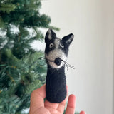 A charming, handcrafted badger felt finger puppet is held by a female hand, set against a soft, blurred background with a subtle Christmas tree ambiance. This unique badger finger puppet, needle-felted from 100% wool, features a black body, a grayish-white face, pointed ears, and delicate whiskers, emphasizing its tactile texture and playful design.