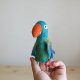 A handmade wool felt blue parrot finger puppet is delicately held up close by a female hand against a bright, neutral backdrop. This handcrafted puppet features a vibrant blend of blue and green wool felt on its body, complemented by a striking orange beak and small wings with subtle shades of blue and yellow, highlighting its tactile texture and playful design.