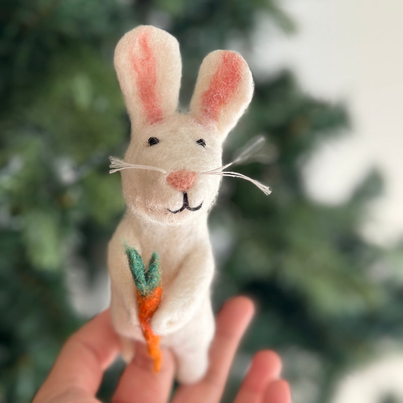 A soft, white handmade felt bunny finger puppet holding a meticulously crafted felt carrot is gently held by a female hand against a blurred Christmas tree backdrop. The bunny features long ears with pink accents, a tiny pink nose, black eyes, and a small black stitched mouth. This charming handcrafted felt toy, made with fair-trade practices, is perfect for imaginative play and storytime.