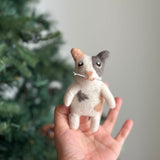 A charming needle felted calico cat finger puppet is gently held in a female hand, set against a soft, neutral background with a hint of a Christmas tree, creating a cozy, playful atmosphere. This handcrafted felt kids toy features distinct patches of peach, white, and grey, including a love spot on its round body. It has tiny arms, perky upright ears, and a unique heart-shaped pink nose.
