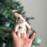 A handmade calico cat felt finger puppet is gently held by a female hand, set against a blurred Christmas tree. This wool felt finger puppet features distinct markings, including a light brown ear, a gray patch near the other ear, and a gray side spot. It has a unique heart-shaped pink nose, white whiskers, and small black eyes, embodying charming craftsmanship.