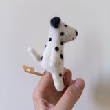 A handmade felt dalmatian dog finger puppet is held up by a female hand against a light background. This unique felt puppet features a white body with characteristic black spots, black ears, and a small attached tag.