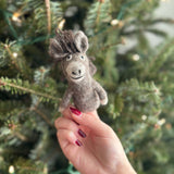 A handmade gray wool felt donkey finger puppet is held in a female hand, interacting playfully with a blurred Christmas tree background adorned with warm lights. This needle felted donkey puppet, crafted in 3D, features a fluffy dark brown mane, small upright ears, and a warm stitched smile, embodying a whimsical holiday character.