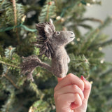 A handmade felt gray donkey finger puppet is held up by a hand, set against a blurred, softly lit Christmas tree. This charming needle-felted wool donkey puppet features a soft, fuzzy texture, a darker wool felt mane and tail, a small stitched smile, and upright ears, all contributing to its three-dimensional design.