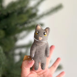 A handcrafted gray cat felt finger puppet is held playfully by a female hand against a blurred background with a glimpse of a Christmas tree. This unique felt toy features small, pointed ears with tan interiors, enchanting yellow big eyes, a tan nose, and white whiskers, ready for imaginative fun.