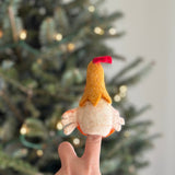 A fair-trade handcrafted felt rooster finger puppet is held on an index finger, set against a blurred Christmas tree background. This unique, vibrant yellow rooster features a red crest and white and orange wings, showcasing its soft, textured felt material.