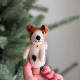 A handmade wool felt Jack Russell terrier dog finger puppet is held by a female hand with red nails, set against a blurred background resembling a Christmas tree. This charming felt finger puppet features a black nose, folded ears, a bell necklace, and a distinctive black love spot on its side, showcasing its meticulous craftsmanship.