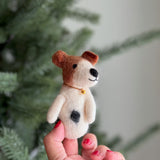 A wool felt Jack Russell Terrier finger puppet is held by a female hand with red nail polish, set against a softly blurred Christmas tree background. This meticulously crafted puppet is mostly white, featuring brown folding ears, a brown patch on its head, and an irresistible smile. A distinctive black 'love spot' adorns its side, complemented by a delicate gold-colored bell necklace around its neck.