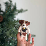 A handmade wool felt Jack Russell terrier finger puppet is held upright in a female hand, set against a blurred Christmas tree. This meticulously crafted dog finger puppet is predominantly white with distinct brown and black markings. It is adorned with a gold bell and a brown heart-shaped spot, offering a unique toy for imaginative play.