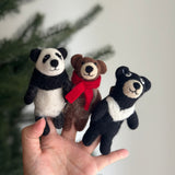 A handmade felt Kung Fu Panda finger puppet is playfully showcased on a female hand against a soft background with a partial view of a Christmas tree. This black and white felted finger puppet features distinct black ears and eye patches, along with a cheerful, smiling expression. Its textured wool felt material highlights the unique craftsmanship of this felt toy for kids.