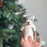 A handmade gray and white felt cat finger puppet, 'Mochi', is presented held upright in the palm of a female hand. This charming handcrafted felt kids toy features a gray head, white body, small round eyes, a brown nose, and delicate white whiskers, set against a blurred, warm background with hints of a Christmas tree.