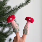 A handmade wool felt mushroom finger puppet is held by a female hand, posed against a soft white backdrop and the green needles of a Christmas pine tree. This charming mushroom puppet features a bright red cap and a cheerful, smiling face, perfect for imaginative play.