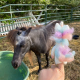 A handcrafted wool felt pastel unicorn finger puppet is held on a finger outdoors. This eco-friendly felt unicorn puppet features a white body and a soft, multi-colored mane in pastel pink, blue, and purple. In the background, a grey pony stands near a green water trough, with a metal gate and straw scattered on the ground.