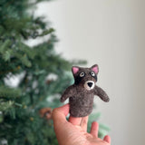 A handmade wool felt raccoon finger puppet is gently presented by a female hand, set against a blurred, festive Christmas tree background. This charming felt finger puppet features a brown body, a black mask around its eyes, pink inner ears, white eyes, and a white muzzle, offering a unique toy for kids' finger play.