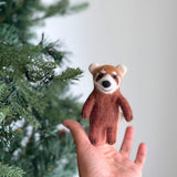 A needle felted red panda finger puppet is playfully displayed against a festive Christmas tree adorned with lights. This unique finger puppet features reddish-brown fur and distinctive white face markings, capturing a charming holiday mood.