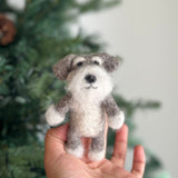 A handmade Schnauzer dog felt finger puppet is held by a female hand, set against the soft, diffused background of a cozy Christmas tree. This unique felt finger puppet features the Schnauzer's distinctive gray and white wool felt beard and bushy eyebrows. Its rounded ears and simple stitched facial features add charm, highlighting the detailed craftsmanship of this handcrafted felt kids toy.