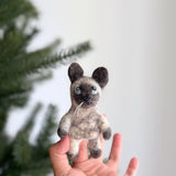 A handcrafted felt Siamese cat finger puppet is held closely in a female hand, playfully set against a blurred Christmas tree background. This charming felt puppet features a grey face with a white section extending from its nose to its chest, complemented by dark grey ears, paws, and tail. Blue eyes and white whiskers add to its adorable feline look. This unique, handcrafted toy is made with ethical and eco-friendly practices.