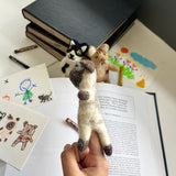 A cream and brown Siamese cat felt finger puppet is held by a hand, presented in a whimsical setting with stacked books, children's drawings, and art supplies in the background. Crafted from plush felt, this unique and handmade finger puppet is an eco-friendly toy. Its fair-trade craftsmanship supports artisans and adds a charming touch to playtime.