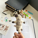 A handmade wool felt Siamese Cat finger puppet is held by a hand, presented in a playful, educational setting with an open book, drawings, and crayons. Two other cat felt finger puppets are softly blurred in the background. This uniquely crafted felt toy, made from soft, textured wool felt by a Fair Trade artisan collective in Nepal, fosters creative play.