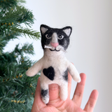 A handcrafted black and white tuxedo cat felt finger puppet is held by a female hand against a festive Christmas tree backdrop. This charming plush felt puppet features a pink nose, white whiskers, and a black heart on its chest, perfect for imaginative play.